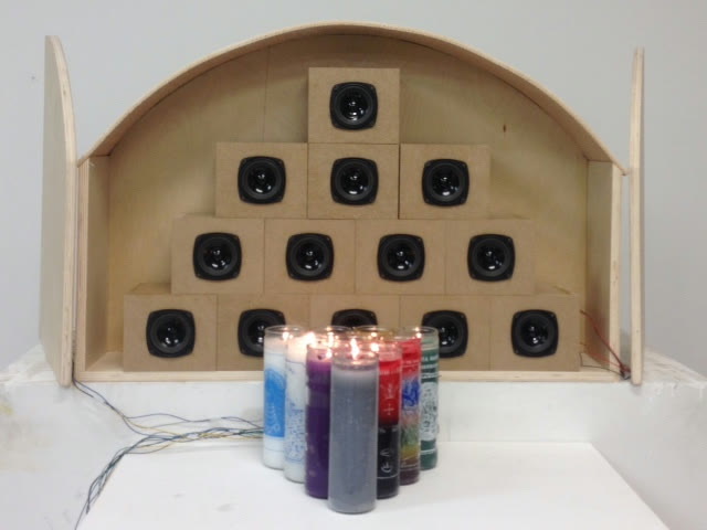 A wooden shrine holding thirteen speakers in a pyramid above a row of lit devotional candles