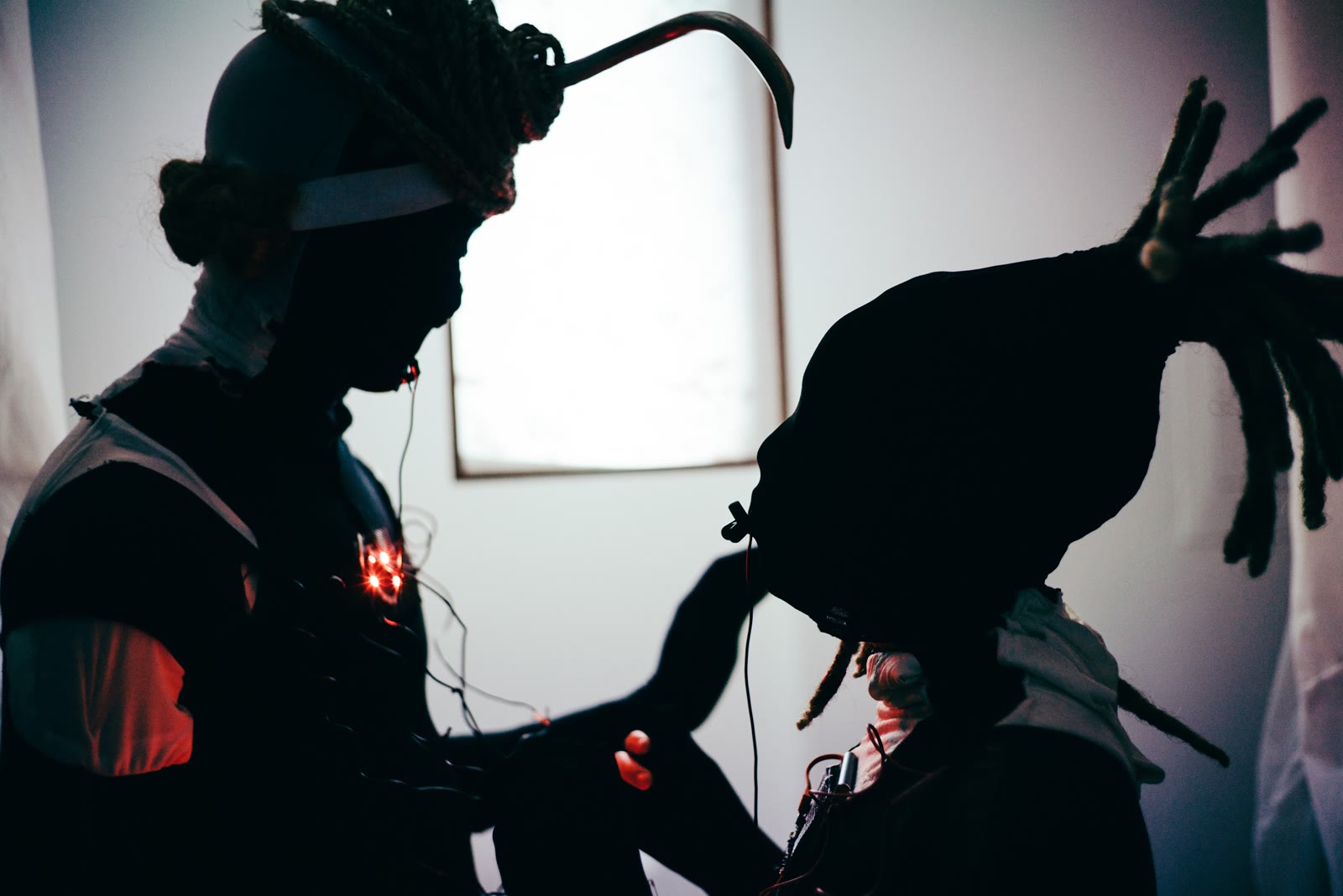 Two silhouetted performers in horned headpieces reaching toward each other, a red light glowing between them
