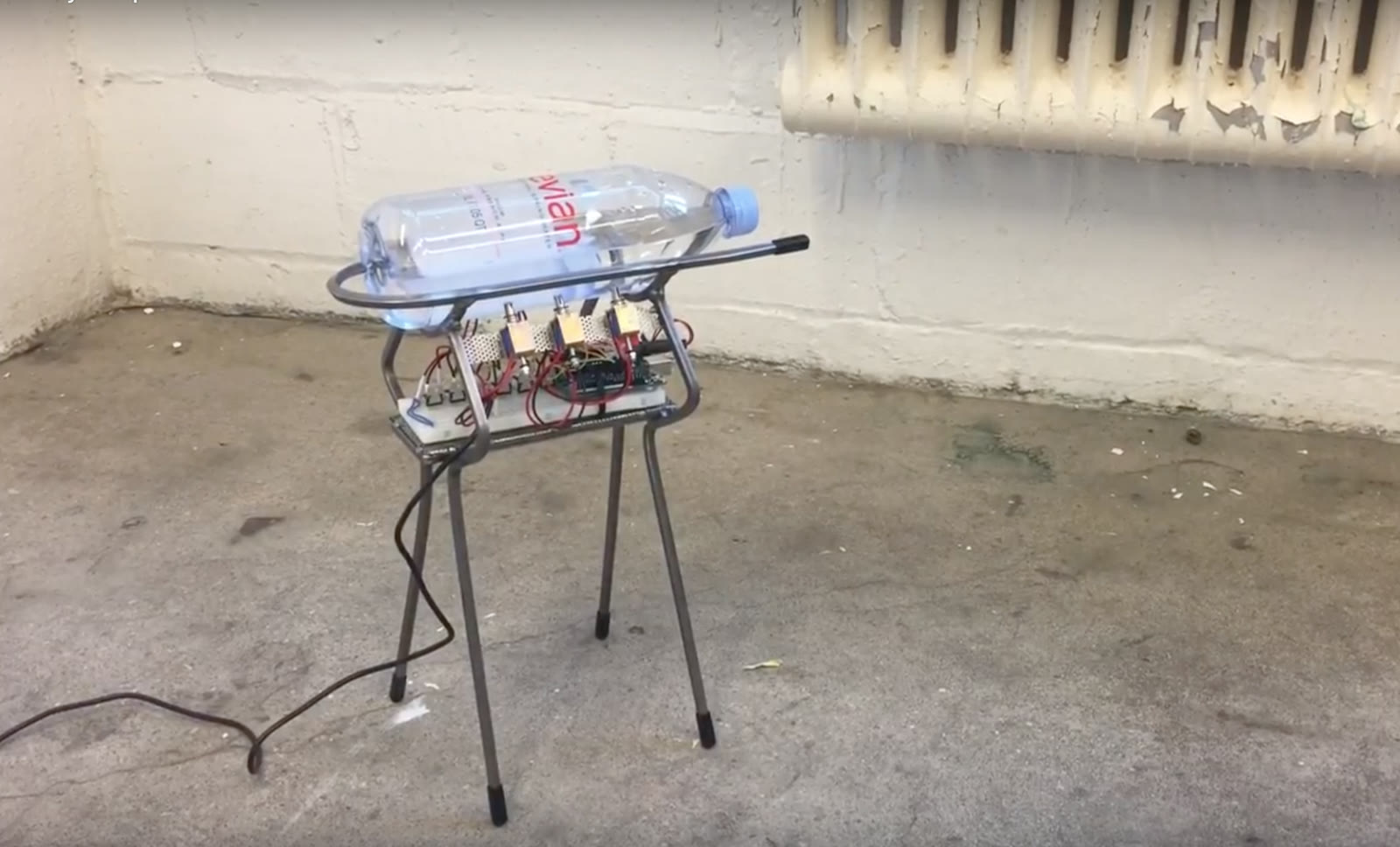 A water bottle resting on a wire stand rigged with electronics against a concrete wall